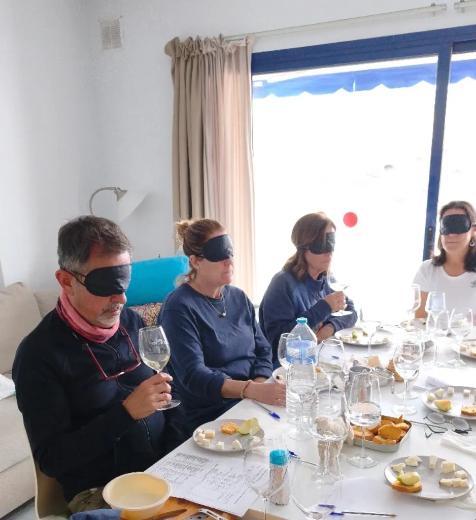 Cuatro asistentes con antifaz catan a ciegas vino blanco y quesos en una mesa junto a una ventana, con platos de queso en tacos, manzana, tostas y fichas de cata.