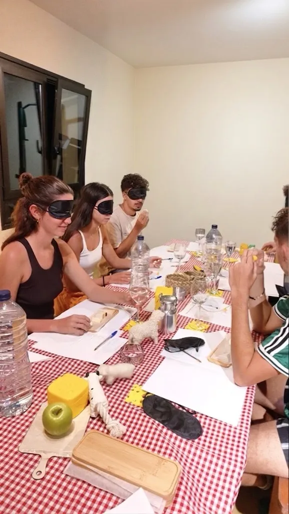 A group of guests wearing blindfolds taste cheese blind around a table with a red checked cloth, with score sheets and glasses of water.