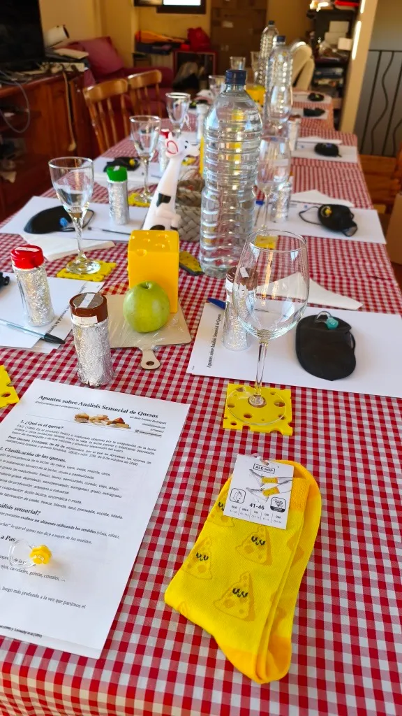 Detail of a tasting table: notes on cheese sensory analysis, aroma jars wrapped in foil, blindfolds, a green apple and glasses.