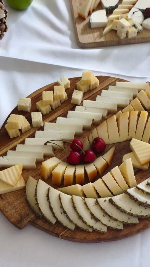 Tabla redonda de madera con quesos en cuñas y tacos dispuestos en abanico, con cerezas en el centro y una manzana verde y una segunda tabla al fondo.